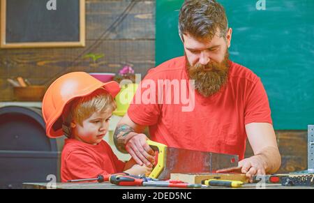 Concetto di paternità. Ragazzo, bambino occupato in casco protettivo imparare ad usare la sega a mano con papà. Padre, genitore con barba che insegna a piccolo figlio a segare con sega a mano affilata, mestieri falegname. Foto Stock