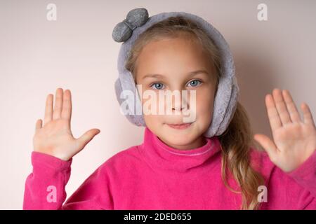 Bambina carina ha sollevato due palme e guarda la fotocamera, è in pelliccia calda cuffie su uno sfondo chiaro Foto Stock