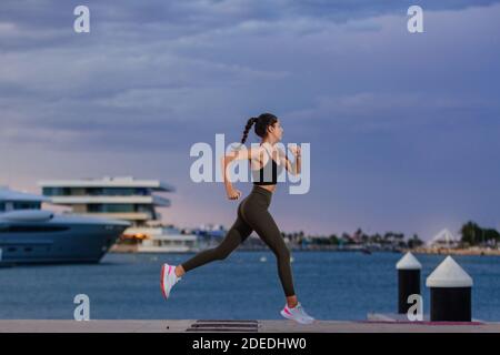 Runner vestito di abbigliamento sportivo corre lungo il mare in un porto con barche ormeggiate. La ragazza è giovane e ha un corpo atletico Foto Stock