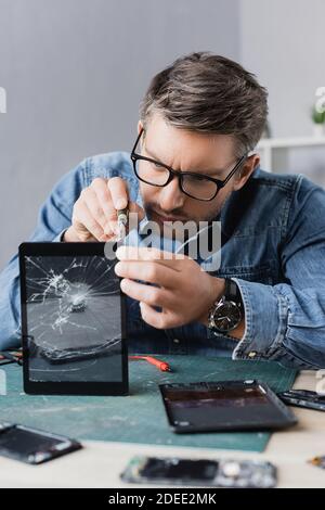 Riparatore mirato con cacciavite che fissa il display di una compressa schiacciata con luogo di lavoro sfocato in primo piano Foto Stock