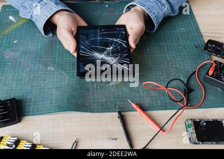 Vista ritagliata del riparatore che tiene in mano un tablet digitale schiacciato sul luogo di lavoro vicino al multimetro e telefoni cellulari smontati Foto Stock