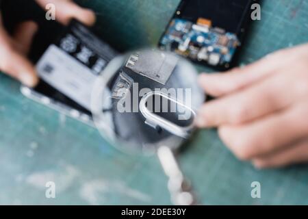 Vista ritagliata della lente di ingrandimento di tenuta del riparatore sulle parti rotte di telefoni cellulari in primo piano sfocato Foto Stock