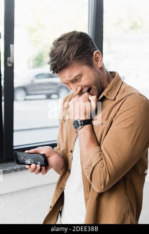 Uomo d'affari spaventato che mordicchiava il pugno mentre si guarda lo smartphone smash con finestra sfocata sullo sfondo Foto Stock