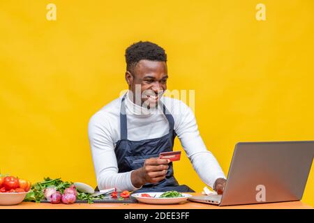 felice chef africano utilizzando il suo computer portatile e tenendo un credito scheda Foto Stock