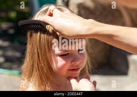 Madre che combatte i capelli delle sue figlie, bambino che viene pettinato dalla sua mamma usando un pettine nero, un primo piano, un ritratto. Donna che fa i capelli felici delle bambine, all'aperto Foto Stock