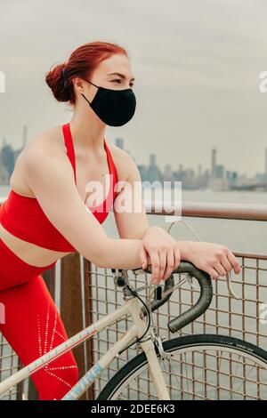 Giovane ciclista femminile che indossa maschera facciale con bicicletta sul lungomare Foto Stock