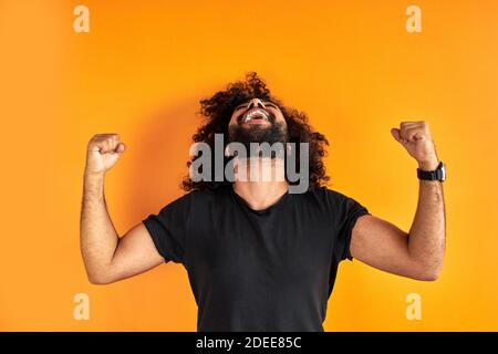 felice uomo bearded celebrare il suo vincitore, sorriso, ragazzo curly brillare con felicità, concetto di vittoria. isolato sfondo arancione Foto Stock