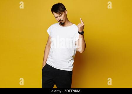 Ragazzo hipster con barba e baffi in bianco tshirt bianca vuota che punta il dito verso l'alto e posa su sfondo giallo, isolato. Spazio di copia per il logo o. Foto Stock