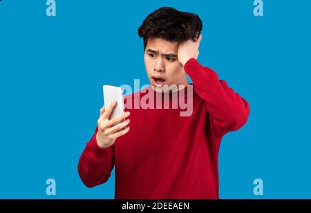 Uomo asiatico sorpreso utilizzando il telefono cellulare sulla parete blu Foto Stock