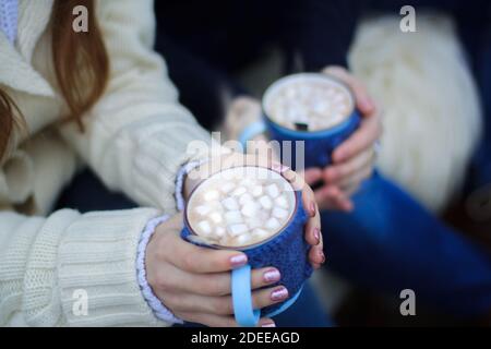 Le mani di una ragazza e di un ragazzo stanno tenendo tazze in maglia blu con cacao e marshmallows bianchi Foto Stock