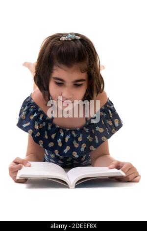 Carino ispanica piccola ragazza che posa sul pavimento e la lettura Un libro - isolato su uno sfondo bianco Foto Stock