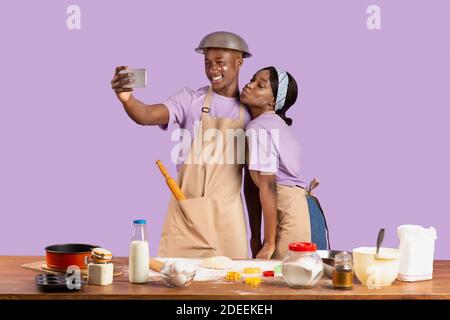 Coppia nera positiva con facce ricoperte di farina che prende selfie mentre cucinando insieme su sfondo viola studio Foto Stock