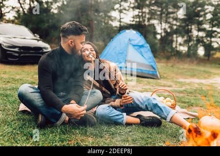 Bella coppia di amanti con arrostire marshmallows in mani spendere il fine settimana in natura con tenda vicino al fuoco Foto Stock