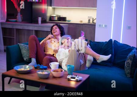 Felice fidanzato e ragazza ridendo dopo aver giocato a videogiochi a casa, joysticks e spuntini su un tavolo. Coppia millenaria che si diverta a passare il tempo Foto Stock