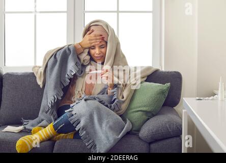 Giovane donna stressata in abiti caldi e coperta che misura il corpo temperatura e sensazione di febbre Foto Stock