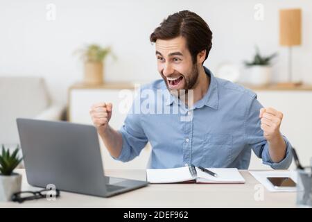 Eccitato giovane uomo d'affari al computer portatile scuotendo i Fists in ufficio Foto Stock