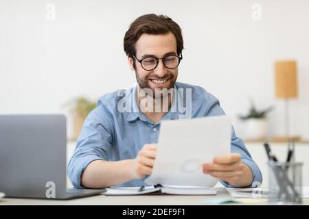 Imprenditore di successo che guarda attraverso i documenti seduti al portatile in ufficio Foto Stock