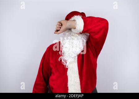 Uomo vestito come Babbo Natale in piedi su isolato sfondo bianco che copre gli occhi con braccio sorridente allegro e divertente. Concetto cieco. Foto Stock