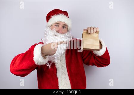 Uomo vestito come Babbo Natale in piedi su isolato sfondo bianco tenendo un sacchetto di carta e puntando il sacchetto Foto Stock
