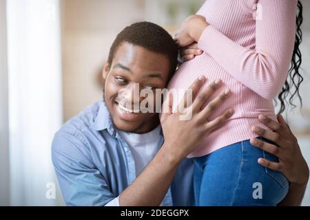 Ragazzo nero premuroso che gli ha messo l'orecchio sulla pancia incinta della ragazza, ascoltando il battito cardiaco del bambino a casa Foto Stock