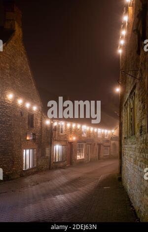 Luci di Natale lungo la strada della chiesa nella nebbia di notte. Stow sul Wold, Cotswolds, Gloucestershire, Inghilterra Foto Stock