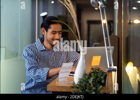 Giovane uomo d'affari asiatico adulto seduto al bar in ufficio con computer portatile. Concetto di lavoro intelligente ovunque. Foto Stock