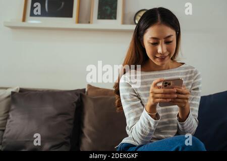 30s giovane donna asiatica adulta che gioca un telefono cellulare si siede sul divano nel soggiorno a casa. Happy donna internet e la tecnologia utente in abiti casual sul divano in camera accogliente Foto Stock