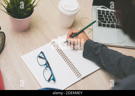 Donna mano che scrive le sue idee creative su notebook vuoto con matita accanto al suo computer portatile su una scrivania di legno con caldo bevanda nella tazza da portare via Foto Stock