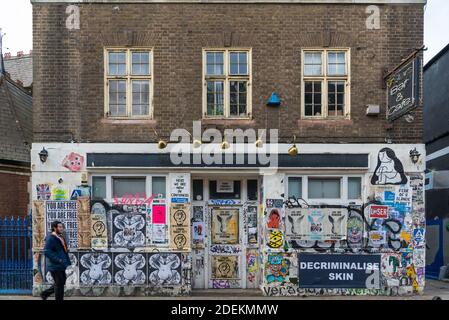 Un uomo solista cammina oltre il 7 Stars Bar and Cafe, a Brick Lane, Spitalfields, Londra. La parte anteriore dell'edificio è coperta da poster e graffiti. Foto Stock
