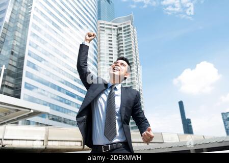 Uomo d'affari riuscito che mostra il successo e la vittoria all'aperto su un soleggiato giorno con il pugno nell'aria e sorridendo Foto Stock