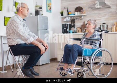 Donna non valida in pensione in sedia a rotelle che ha una conversazione con vecchio marito anziano in cucina. Anziano che parla con la moglie. Vivere con persone disabili con disabilità motorie Foto Stock