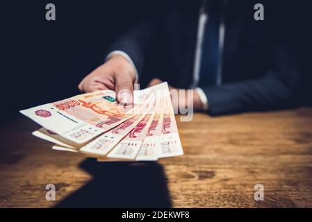 Uomo d'affari che dà denaro, banconote ruble russe, sulla sua scrivania in un ufficio oscuro - concetto di corruzione e corruzione Foto Stock