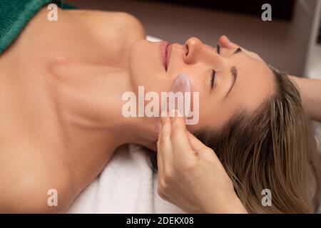 Giovane e bella donna durante il massaggio cinese tradizionale - Gua Sha. Primo piano. Trattamenti di bellezza nel salone SPA. Strumenti di bellezza in mano a bea Foto Stock