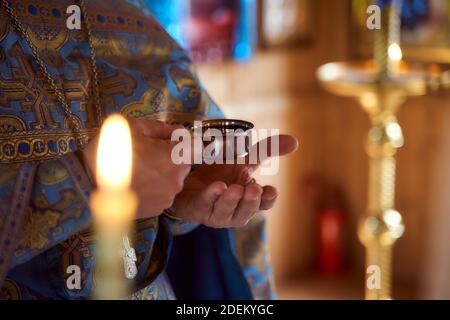 Le mani del sacerdote tengono una piccola ciotola di vino nell'ambiente della chiesa. Candele sullo sfondo Foto Stock