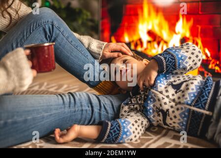 Mamma e figlio piccolo sono seduti accanto al camino, che è raffigurato sullo schermo TV e divertirsi. Il concetto di creare un'accogliente atmosfera invernale Foto Stock