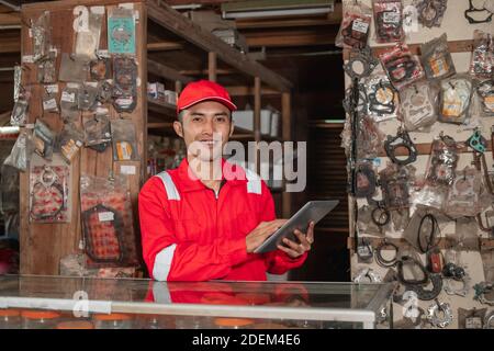 Mechanic sorridente che indossa un auricolare e un cappello utilizzando il supporto del tablet digitale nel negozio di ricambi per autoveicoli Foto Stock