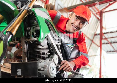 Un mechanic sorridente che indossa un auricolare e un cappello utilizzando una chiave a. serrare il bullone della motocicletta nel garage Foto Stock