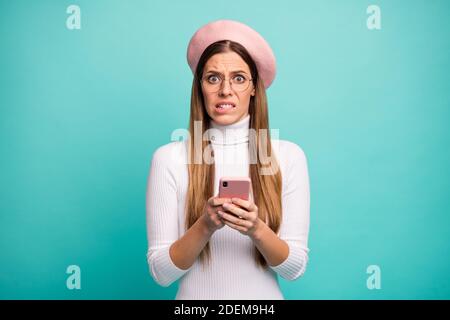 Foto di bella terrorizzata signora tenere telefono leggere nuovo post commenti negativi mordere le labbra usura specifiche moderno berretto rosa dolcevita bianco isolato Foto Stock