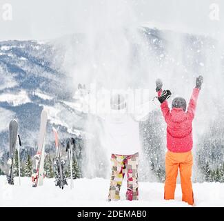 Vista posteriore di giovani coppie di turisti che giocano nella neve contro paesaggi ipnotici sullo sfondo. Abiti vivaci e colorati sulla neve bianca. Momenti indimenticabili. Concetto di attività sportive invernali, divertimento Foto Stock