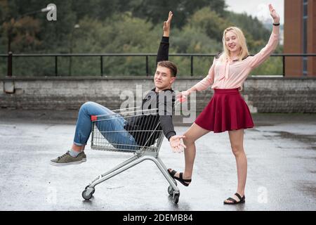 Giovane coppia che si diverte sul carrello dello shopping. Felice donna che spinge il carrello con il suo ragazzo dentro in strada Foto Stock