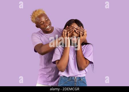 Indovinate chi. Giocoso uomo nero sorprendente la sua ragazza, coprendo gli occhi da dietro sullo sfondo viola studio Foto Stock