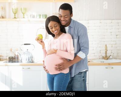 Ritratto di ragazzo nero amorevole abbracciando la sua bella moglie in attesa con mela in cucina Foto Stock