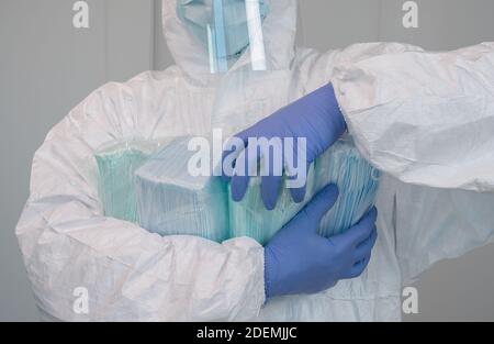 Un infermiere in un vestito protettivo ha diverse confezioni di nuove maschere e sta raccogliendone una. L'operatore sanitario indossa guanti e una maschera facciale per prevenire la corona Foto Stock