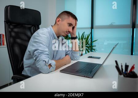 Stanco o stressato giovane uomo d'affari seduto di fronte al computer in ufficio. Imprenditore infelice sul posto di lavoro. Malsano esausto uomo caucasico havin Foto Stock