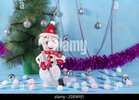 Un pupazzo di neve allegro ma solitario in un vestito rosso con un bastone da hockey si trova accanto a un albero verde una nuova tappa blu dell'anno Foto Stock