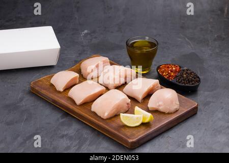 Pollo crudo tenero fritto senza pelle disposto su legno tavole e guarnite con olio, fette di limone, fiocchi freddi e pepe nero su sfondo grigio Foto Stock