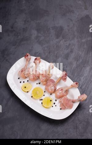 Pollo crudo lollipop, sei pezzi di pollo lollipop disposti su un servire piatto bianco e guarnito con fette di limone e nero pepe su consistenza grigia Foto Stock