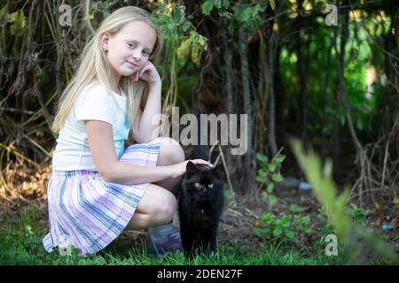 Bambina bionda gioca con un gatto nero su uno sfondo di cespugli verdi. Foto Stock