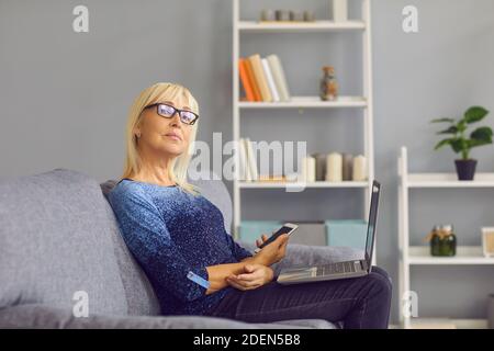 Vista laterale di una donna anziana matura che effettua telefonate e controlla le chat di lavoro sul suo computer portatile. Foto Stock