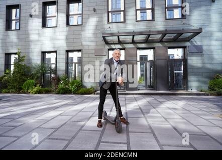 Andare avanti. Foto a tutta lunghezza di un uomo d'affari maturo ed elegante che guarda la fotocamera mentre si trova in piedi con uno scooter elettrico vicino a un moderno edificio d'ufficio Foto Stock